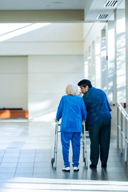 Companion assisting an elderly woman with a walker in a hospital corridor